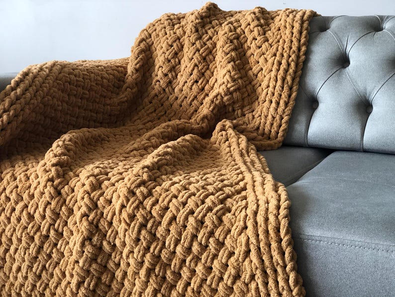 May include: A chunky knit throw blanket in a warm brown color. The blanket is made with a thick yarn and has a textured weave. It is draped over a gray couch.