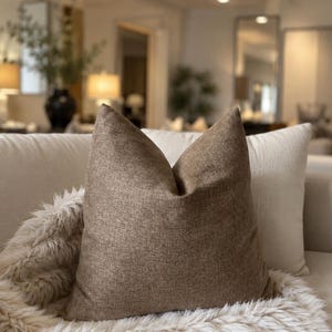 May include: A square throw pillow featuring a textured, medium-brown fabric with a subtle woven pattern. The pillow is positioned upright with a distinct center crease at the top, resting against a larger, plain off-white pillow on a sofa.