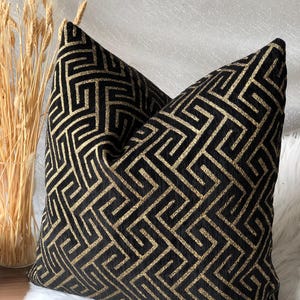 May include: Two decorative throw pillows with a black and gold geometric pattern. The pillows have a textured appearance and are placed on a white, fluffy surface. The design features a repeating maze-like pattern.