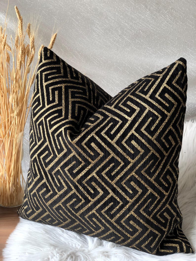 May include: Two decorative throw pillows with a black and gold geometric pattern. The pillows have a textured appearance and are placed on a white, fluffy surface. The design features a repeating maze-like pattern.