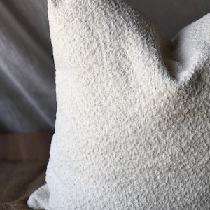 May include: A square throw pillow featuring a textured, off-white boucl&eacute; fabric cover. The material has a distinct nubby, looped surface that creates a dimensional appearance across the entire front face of the pillow, which is filled to a firm shape.