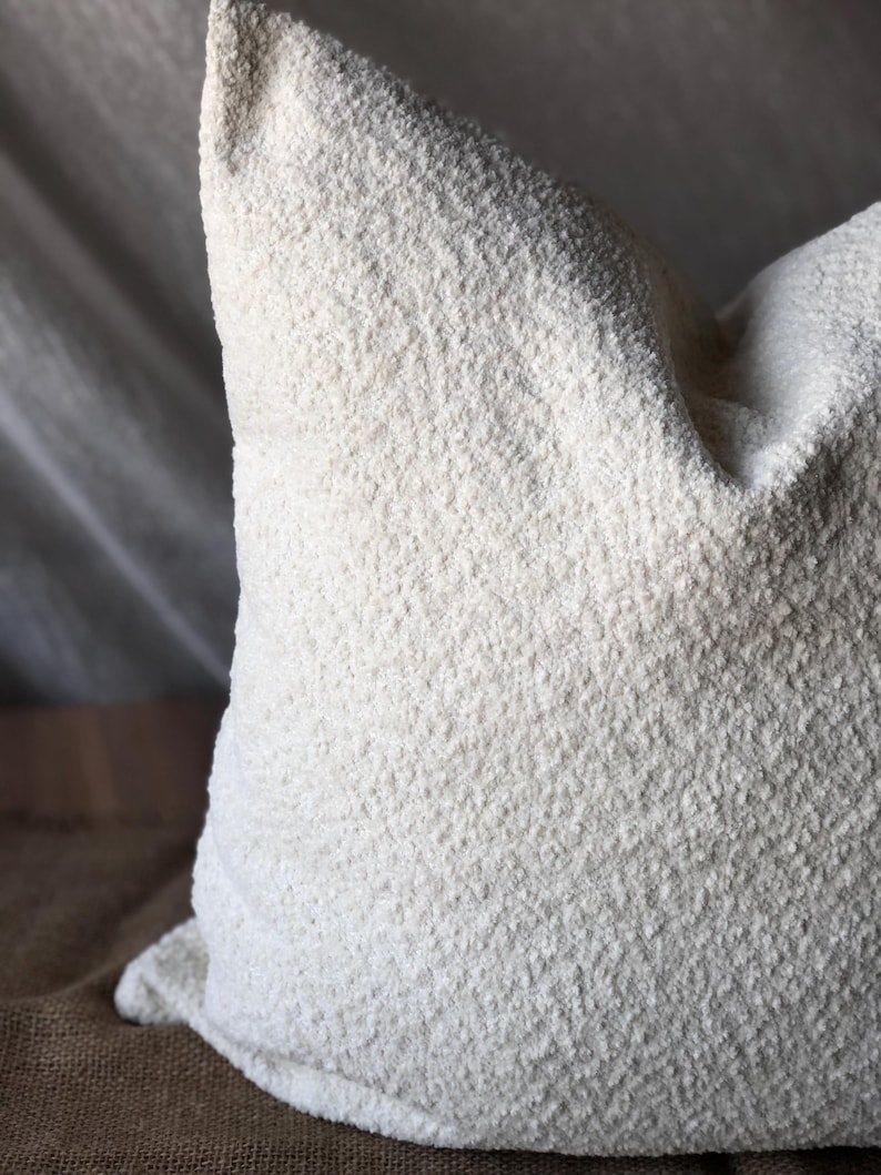 May include: A square throw pillow featuring a textured, off-white boucl&eacute; fabric cover. The material has a distinct nubby, looped surface that creates a dimensional appearance across the entire front face of the pillow, which is filled to a firm shape.