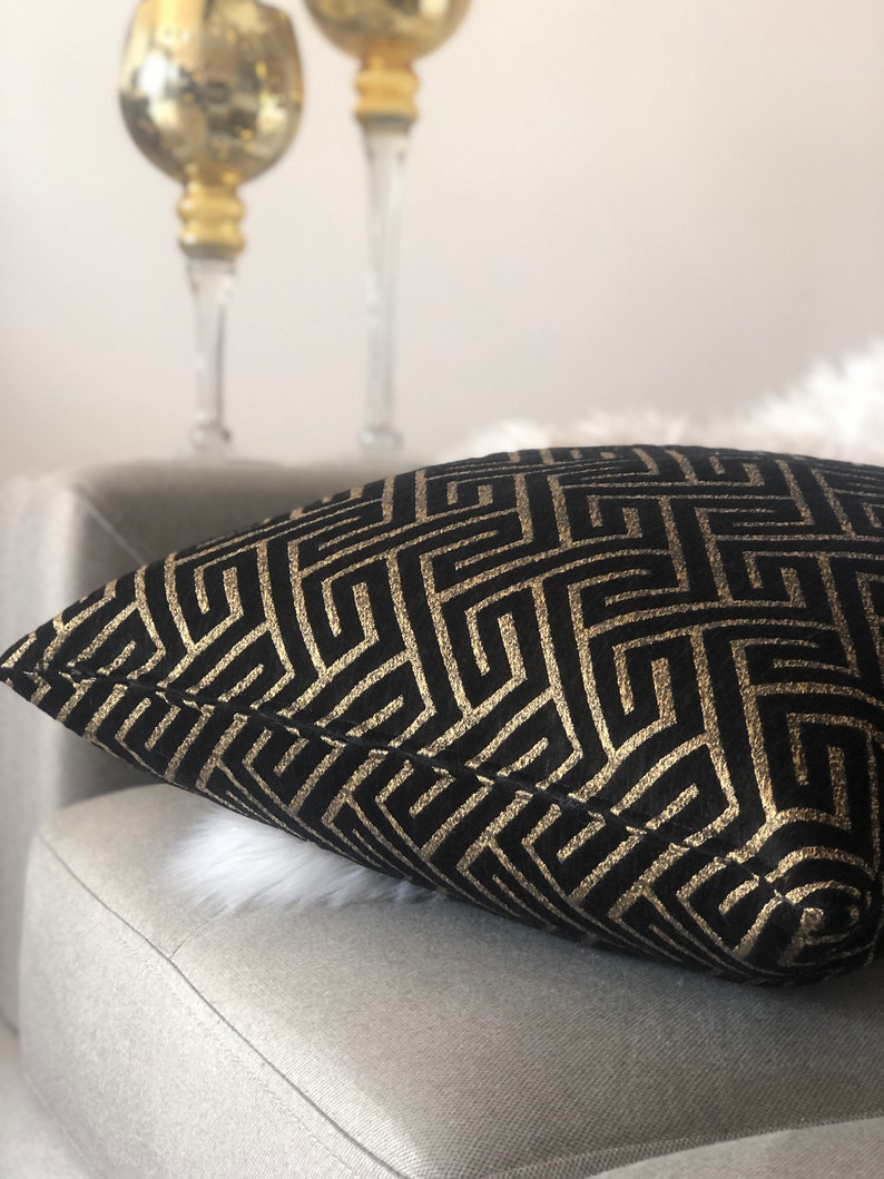 May include: Black and gold geometric patterned throw pillow with a subtle sheen. The pillow is on a light gray couch.