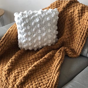 May include: A white knitted throw pillow with a textured bobble design sits on top of a mustard yellow knitted throw blanket. The blanket is folded and draped over a gray couch.
