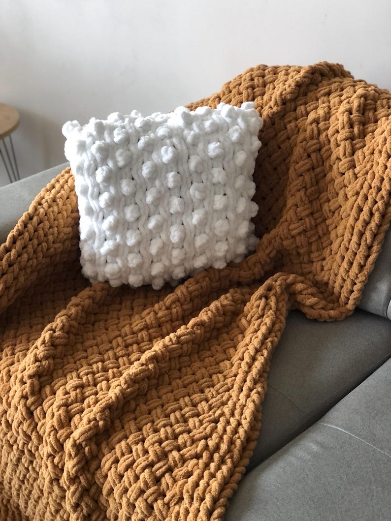 May include: A white knitted throw pillow with a textured bobble design sits on top of a mustard yellow knitted throw blanket. The blanket is folded and draped over a gray couch.