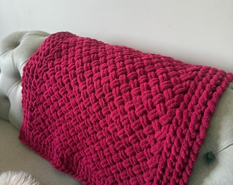 May include: A vibrant, hand-knitted blanket in a rich magenta hue. The blanket features a chunky, woven texture, creating a cozy and inviting appearance. The blanket is displayed on a gray tufted sofa, with a white faux fur throw visible.