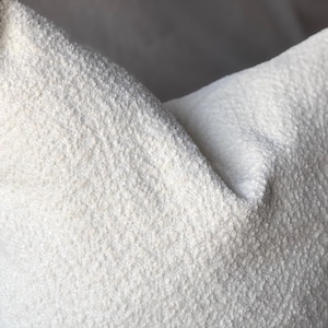 May include: A square throw pillow featuring a textured, off-white boucl&eacute; fabric. The surface of the pillow has a distinct, nubby, and looped appearance, creating a tactile and dimensional look across the entire front face of the cushion.