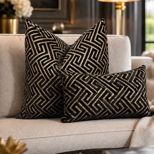 Può includere: Due cuscini decorativi con base in velluto nero e motivo geometrico a greca dorata metallizzata. Un cuscino è quadrato e l'altro è rettangolare, entrambi presentano lo stesso intricato design a linee intrecciate che si ripetono.