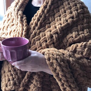 May include: A person wearing a brown chunky knit blanket with a pink mug in their hands.
