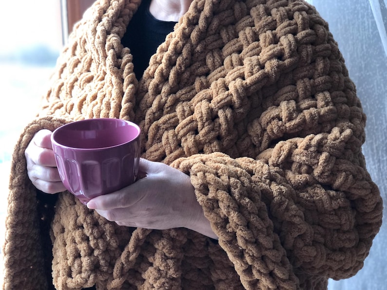 May include: A person wearing a brown chunky knit blanket with a pink mug in their hands.