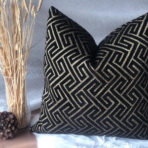 May include: A decorative throw pillow with a black and gold geometric pattern. The pillow is placed on a white, fluffy surface. A clear vase with dried wheat stalks and two pinecones are in the background.