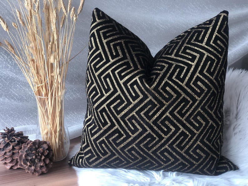 May include: A decorative throw pillow with a black and gold geometric pattern. The pillow is placed on a white, fluffy surface. A clear vase with dried wheat stalks and two pinecones are in the background.