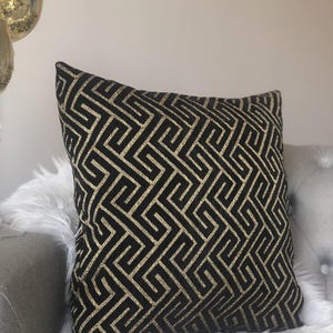 May include: Black decorative throw pillow with a gold geometric pattern. The pillow is square-shaped and rests on a gray surface with white faux fur. The gold pattern creates a striking contrast against the black fabric.
