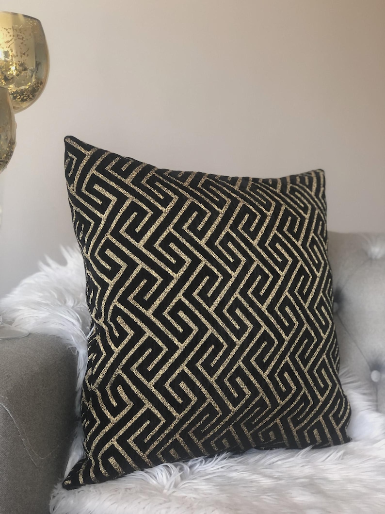 May include: Black decorative throw pillow with a gold geometric pattern. The pillow is square-shaped and rests on a gray surface with white faux fur. The gold pattern creates a striking contrast against the black fabric.