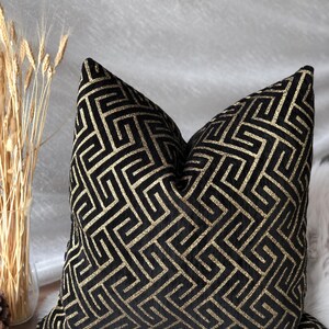 May include: Black decorative throw pillow with a gold geometric pattern. The pillow has a square shape and is made of a soft, textured fabric. The gold pattern is a repeating maze-like design. The pillow is placed on a white surface.