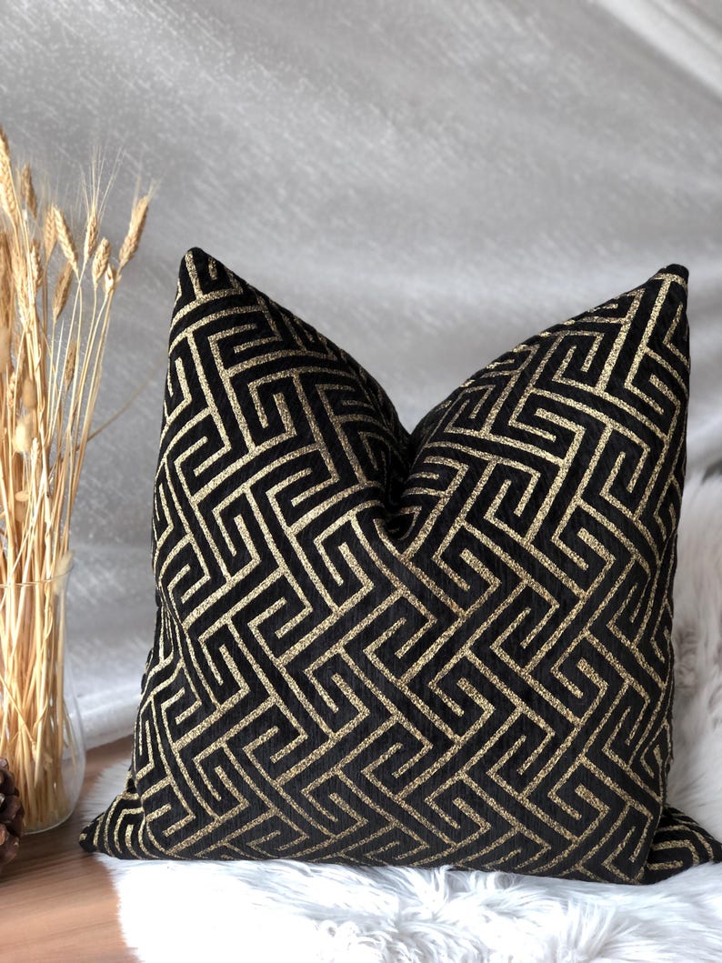 May include: Black decorative throw pillow with a gold geometric pattern. The pillow has a square shape and is made of a soft, textured fabric. The gold pattern is a repeating maze-like design. The pillow is placed on a white surface.