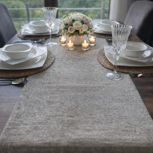 Könnte beinhalten: Ein gedeckter Esstisch für eine Mahlzeit mit weißen Tellern, Schalen und Kristallweingläsern. Ein Blumengesteck und brennende Kerzen sorgen für Ambiente. Ein Tischläufer in neutralen Tönen bedeckt den dunklen Holztisch.
