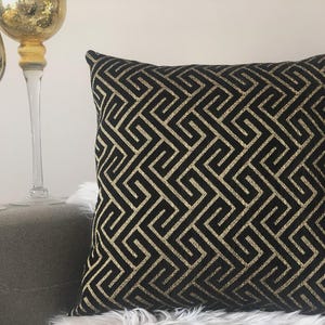 May include: Square decorative pillow with a black background and a gold geometric pattern. The pillow is placed on a gray surface with two gold-colored decorative glass pieces in the background. The pillow has a textured appearance.