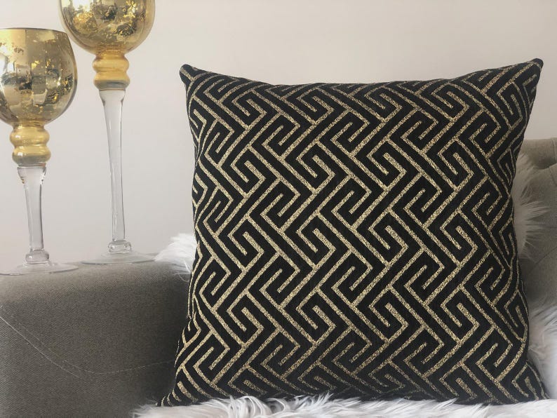 May include: Square decorative pillow with a black background and a gold geometric pattern. The pillow is placed on a gray surface with two gold-colored decorative glass pieces in the background. The pillow has a textured appearance.