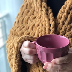 May include: A person wearing a chunky knit mustard yellow cardigan holds a pink ceramic mug with a fluted design.