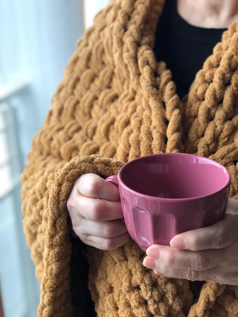 May include: A person wearing a chunky knit mustard yellow cardigan holds a pink ceramic mug with a fluted design.