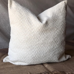 May include: A square throw pillow featuring a textured, off-white boucl&eacute; fabric surface. The pillow has a soft, nubby appearance with a consistent cream-colored finish across the entire front face, showcasing a dense, looped textile construction.