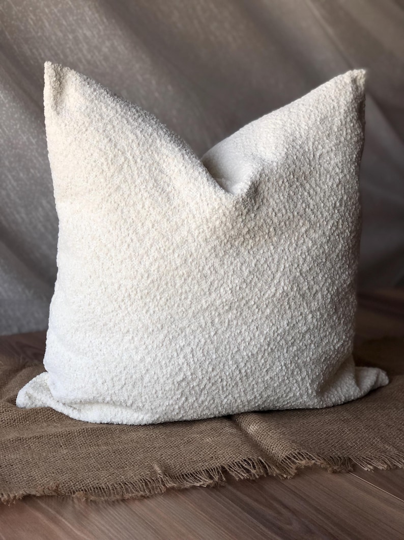 May include: A square throw pillow featuring a textured, off-white boucl&eacute; fabric surface. The pillow has a soft, nubby appearance with a consistent cream-colored finish across the entire front face, showcasing a dense, looped textile construction.