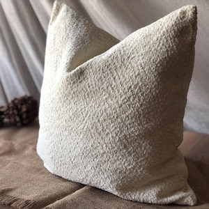 May include: A square throw pillow featuring a textured, off-white, boucl&eacute;-style fabric surface. The pillow has a dense, nubby appearance with a consistent cream color throughout, presenting a soft, voluminous shape with defined corners and edges.