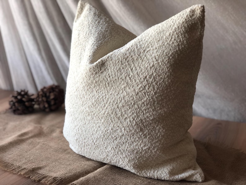May include: A square throw pillow featuring a textured, off-white, boucl&eacute;-style fabric surface. The pillow has a dense, nubby appearance with a consistent cream color throughout, presenting a soft, voluminous shape with defined corners and edges.
