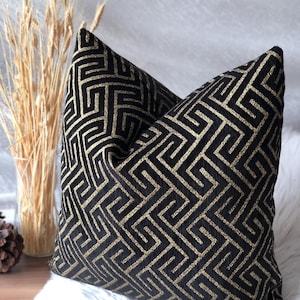 May include: Two decorative throw pillows with a black and gold geometric pattern. The pillows are square-shaped and rest on a white, fluffy surface. A vase of wheat stalks is visible in the background.