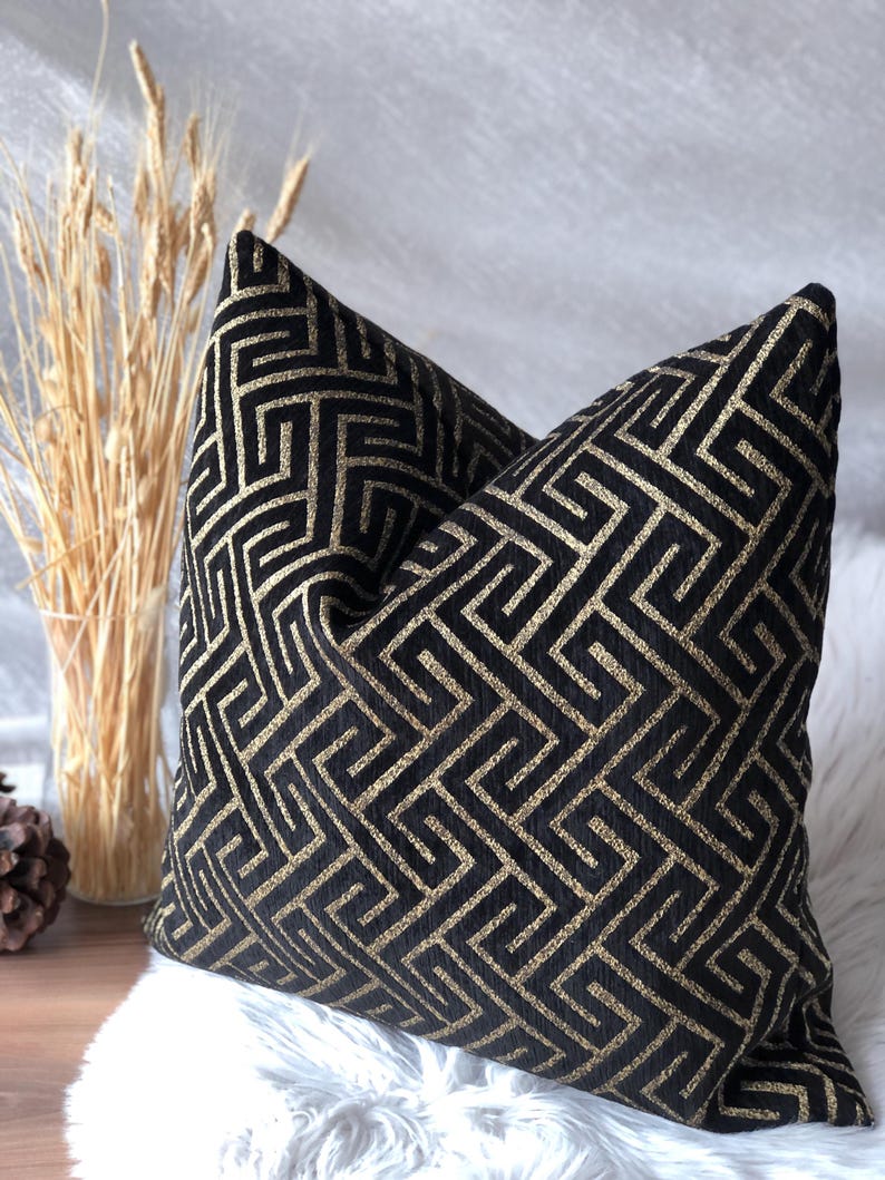 May include: Two decorative throw pillows with a black and gold geometric pattern. The pillows are square-shaped and rest on a white, fluffy surface. A vase of wheat stalks is visible in the background.