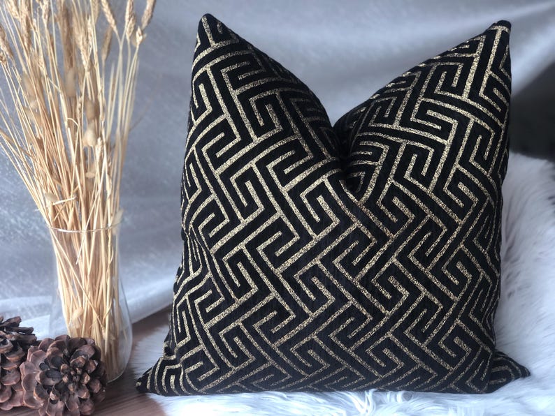 May include: Black decorative throw pillow with a gold geometric pattern. The pillow has a soft, textured appearance and is placed on a white surface. The pillow is a square shape and is a decorative home accent.