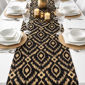 May include: A dining table set for a formal occasion. The table features a black and gold patterned runner, white tableware, and crystal glasses. A floral display and candles enhance the setting.
