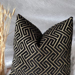 May include: A decorative throw pillow with a black background and a gold geometric pattern. The pillow is square-shaped and has a soft, textured appearance. The design features a repeating Greek key motif.