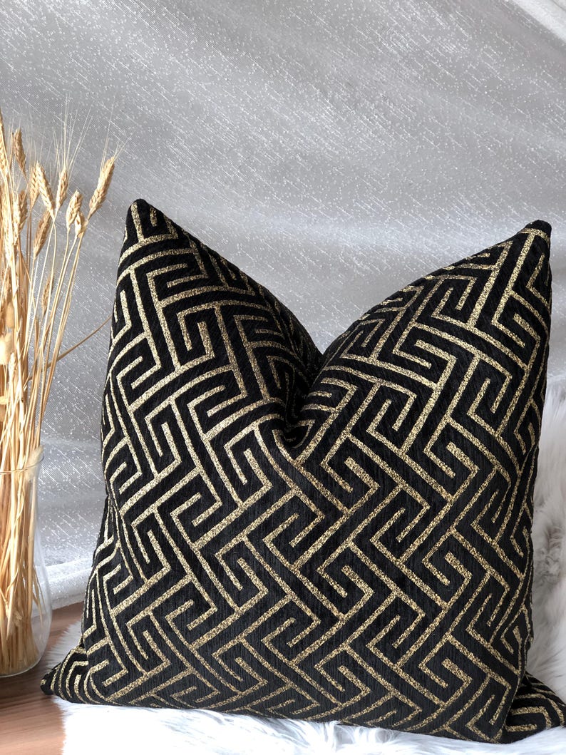 May include: A decorative throw pillow with a black background and a gold geometric pattern. The pillow is square-shaped and has a soft, textured appearance. The design features a repeating Greek key motif.