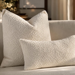 May include: Two off-white throw pillows with a textured, curly, fleece-like fabric surface. One pillow is square-shaped and positioned behind a smaller, rectangular-shaped pillow. Both items feature a consistent, uniform cream-colored finish.