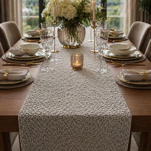 May include: A dining table set for a special occasion, featuring a textured, neutral-coloured table runner. The table is set with fine china, gold cutlery, and a floral arrangement. Glass candle holders and a vase complete the elegant setting.
