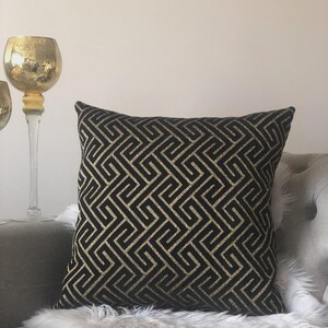 May include: Black and gold geometric patterned throw pillow. The square pillow features a repeating maze-like design in gold against a black background. The pillow is placed on a gray couch with a white faux fur throw.