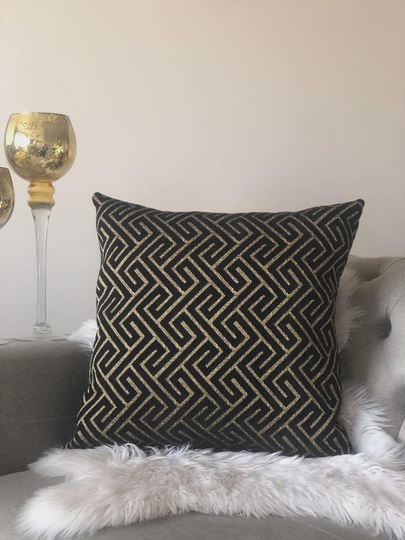 May include: Black and gold geometric patterned throw pillow. The square pillow features a repeating maze-like design in gold against a black background. The pillow is placed on a gray couch with a white faux fur throw.