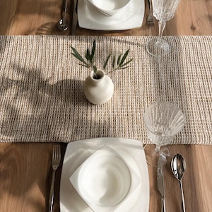 May include: A table setting featuring two square white plates with matching bowls, silver cutlery, and clear ribbed glassware. A beige woven table runner sits in the centre with a small off-white ceramic vase containing green olive branches.