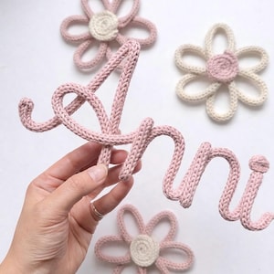 Könnte beinhalten: Eine pinkfarbene Strickkordel in Form des kursiven Namens "Anni" zusammen mit drei gehäkelten Blumendekorationen. Zwei Blumen haben pinke Blütenblätter mit einem cremefarbenen Zentrum, eine Blume hat cremefarbene Blütenblätter mit einem pinken Zentrum.