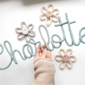 Könnte beinhalten: Ein hellblaues gestricktes Namensschild "Charlotte" mit mehreren gehäkelten Blumenakzenten in neutralen Farben. Die Blumen sind in Creme-, Grau- und Rosatönen gehalten. Der Name ist in Schreibschrift.