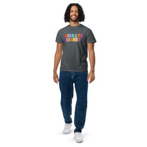 Pode incluir: Uma camiseta cinza escura com o texto "VIBRATE HIGHER" em cores do arco-íris.