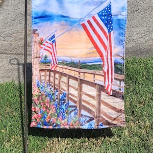 May include: A garden flag featuring a watercolor painting of the American flag waving over a wooden bridge, with colorful flowers in the foreground. The sky is blue with clouds, and the flag is attached to a black metal stake.
