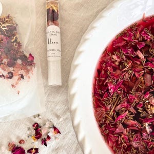 Pode incluir: Uma vista de cima mostra um prato de cerâmica branco cheio de líquido vermelho e pétalas de flores. Um frasco de vidro com rolha de cortiça contém ingredientes para banho. Uma bolsa transparente de sais de banho e pétalas soltas também são visíveis. O frasco diz "Organic Bath Soak Bloom Flower + Still 1 oz."