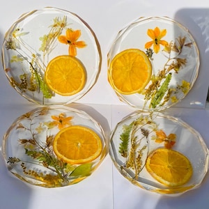 Könnte beinhalten: Vier runde, transparente Harz-Untersetzer mit getrockneten Blumen und Orangenscheiben. Die Untersetzer haben einen geometrischen, facettierten Rand.