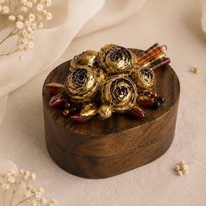 Könnte beinhalten: Eine dunkelbraune, ovale Holzschatulle mit einer dekorativen Verzierung auf dem Deckel. Das Arrangement besteht aus mehreren metallisch goldfarbenen Blüten, kleinen dunkelroten Blättern und drei dünnen, gestreiften braun-beigen Stäbchen.