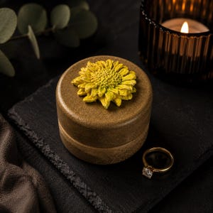 May include: A round, brown, textured container with a sculpted yellow sunflower attached to the lid. A gold-toned ring with a clear stone sits next to the container. The container has a smooth, matte finish and a cylindrical shape with a lid.