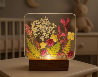May include: A clear acrylic lamp base with a wooden stand, featuring pressed flowers and ferns in shades of pink, yellow, and green. The lamp is illuminated from behind, creating a warm glow.