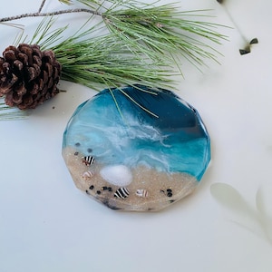 Puede incluir: Un posavasos redondo de resina con temática oceánica con una escena de playa. El posavasos presenta capas de resina azul y blanca para imitar las olas, con arena, conchas marinas y pequeñas piedras negras. Una piña y una ramita de agujas de pino están en el fondo.